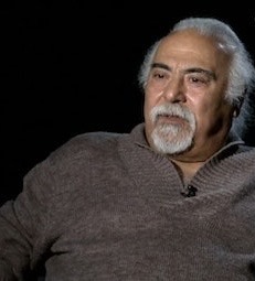 A man with white hair and beard sitting in front of a black background.